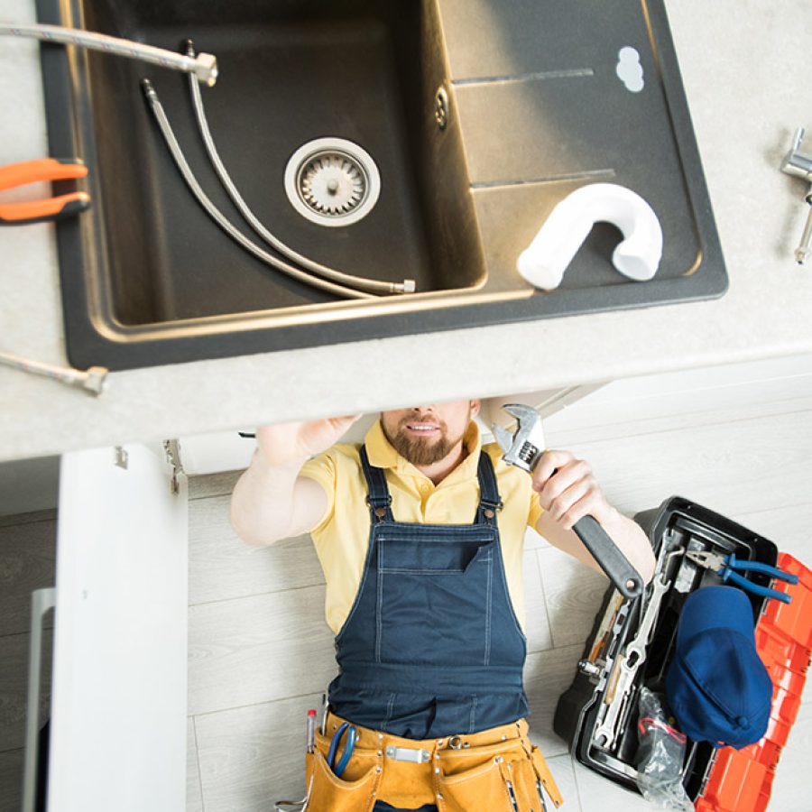 Directly above view of smiling handsome young plumber with beard lying on floor and using wrench while repairing water pipe in kitchen